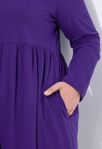 Lila Langarmkleid mit geraffter Taille und Seitentasche. Glatte Stofftextur mit lockerer Passform und rundem Ausschnitt.