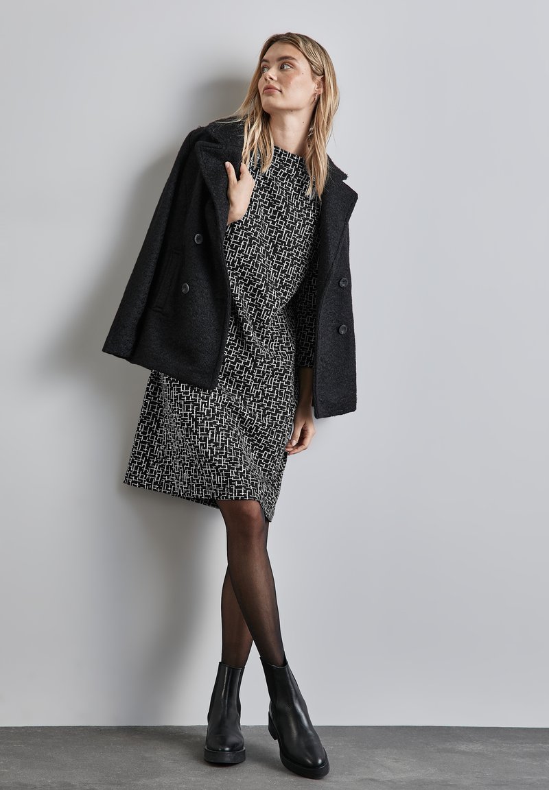 Schwarzer, doppelt geknöpfter Mantel über einem gemusterten schwarz-weißen Kleid, mit schwarzen Strumpfhosen und Stiefeletten, vor einem neutralen Hintergrund.
