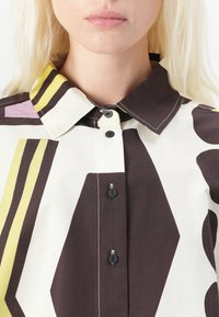 Chemise à boutonner avec une base blanche ornée de grandes formes abstraites en marron, jaune et rose. Boutons noirs et col structuré.