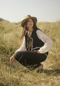 Gilet noir avec des bordures blanches sur une blouse blanche ; chapeau beige à large bord ; pantalons noirs longs ; collier de perles stratifiées ; cadre naturel avec de l'herbe.