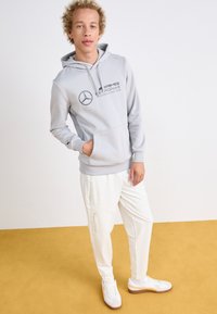 Lichtgrijze hoodie met een voorkwauwtje en zwart logo, gecombineerd met witte tapered broek met ritsen en witte sneakers met bruine zolen.