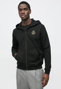 Czarna bluza z kapturem na zamek, wykonana z miękkiego materiału, z przednią kieszenią i złotym logo na lewej piersi. Gładka faktura z prążkowanymi mankietami.