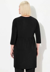 Musta tunikamekko, jossa on kolmennes käsi, kerätty vyötärö ja suora malli, valmistettu pehmeästä ja joustavasta kankaasta.