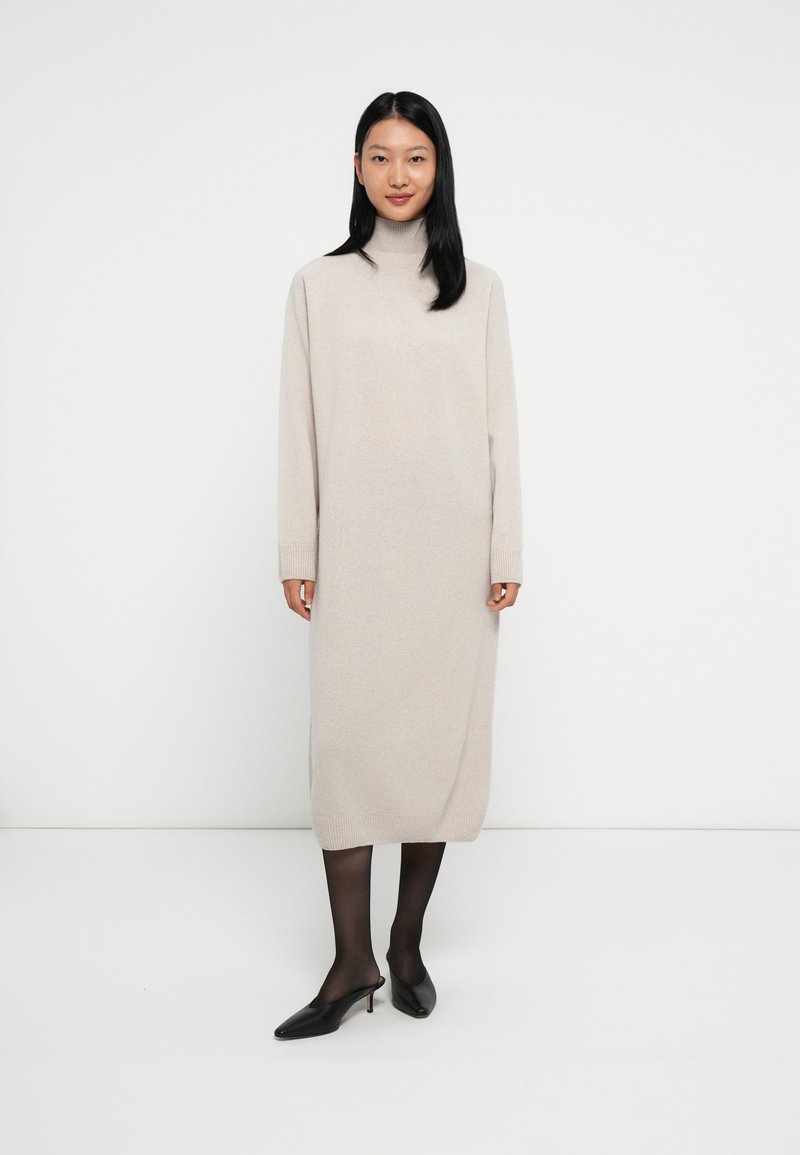 Vrouw in een beige turtleneck midi-jurk, zwarte sheer panty's en zwarte puntige kitten heel mules voor een witte achtergrond.