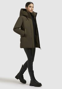 Olivgrüne Steppjacke mit Kapuze, mit einem Reißverschluss vorne, seitlichen Taschen und einem leicht lockeren Schnitt, kombiniert mit schwarzen Leggings und Stiefeln.