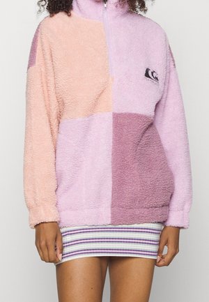 Femme portant une veste polaire à fermeture éclair quartée avec des blocs de couleur rose et violet sur une jupe courte rayée.