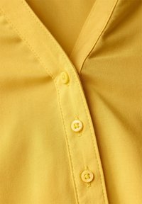 Primo piano di un tessuto di camicia gialla che mostra tre bottoni abbinati e dettagli di cucitura lungo la patta dei bottoni.