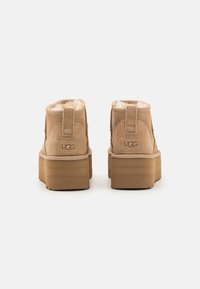 Tanfarbene Wildleder-Stiefeletten mit weichem Futter, flacher Plateausohle und verstärktem Fersenschlaufe; UGG-Logo auf der Rückseite.