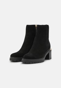 Bottines noires en daim avec un talon épais et une semelle texturée, dotées d'une fermeture éclair dorée et d'un design élégant et moderne.