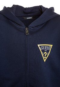 Haut à capuche bleu marine avec fermeture éclair, fabriqué en tissu côtelé. Présente un patch de logo triangulaire jaune avec "GUESS" et un point d'interrogation.