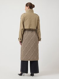 Khaki krótkogolfowy płaszcz trench z wysokim kołnierzem, połączony z długim, pikowanym beżowym płaszczem puchowym. Modelka nosi ciemne dżinsy o szerokich nogawkach i czarne szpilki.