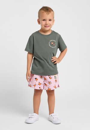 Donkergroene katoenen t-shirt met een witte grafische print. Roze shorts met oranje vogelpatronen. Witte instappers.