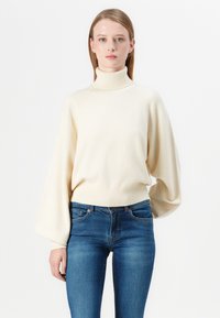 Kremasto turtleneck pulover z ohlapnim krojem in balonskimi rokavi, v kombinaciji s prilegajočimi modrimi kavbojkami. Diskretna rebrasta tekstura na robovih in manšetah.