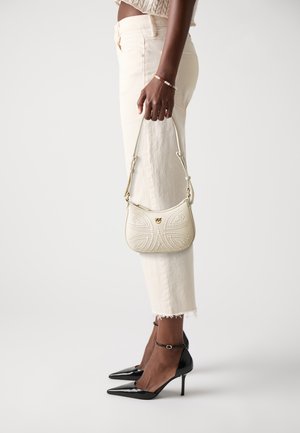 Woman in cream cropped jeans and black heels holding a small cream leather handbag with decorative stitching.