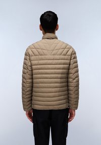 Beige pufferjakke med kviltet design og høj krave, prydet med syede vandrette og diagonale linjer, båret over sorte bukser.