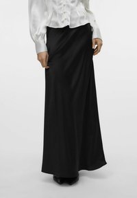 Zwarte, lange rok van gladde stof met een wikkeldesign. G gedragen met een witte blouse met knopen en zwarte schoenen.