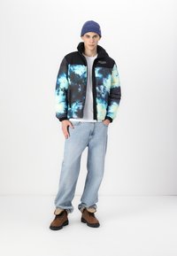 Polstret jakke med et sort og blåt tie-dye mønster, der har lynlåslukning og elastiske manchetter. Kombineret med lyseblå jeans og brune støvler.