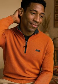 Oranje ritsvest met een zwarte rits en logo-detail. Geribbelde mouwen en onderrand, textuurstof, gedragen over een donker shirt.