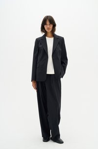 Black tailored blazer with subtle shoulder padding, paired with loose black trousers. Underneath, a plain white shirt. Smooth fabric texture.