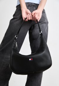 Bolso hobo de piel sintética negra con forma curva, que presenta un zipper plateado y un pequeño logo multicolor. Textura suave y correa ajustable.
