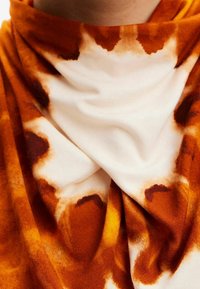 Wielokolorowy szalik z wzorem tie-dye w odcieniach pomarańczowym, brązowym i białym. Materiał wydaje się miękki i swobodnie opada.