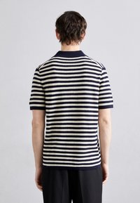 Muž otočený zády, oblečený v tričku s krátkým rukávem v námořnické modré a bílé vodorovné pruhy a černých kalhotách na jednobarevném světlém šedém pozadí.