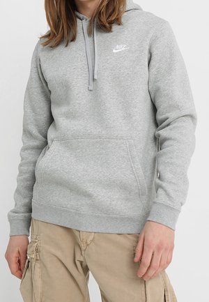Osoba nosząca jasnoszarą bluzę z kapturem Nike z przednią kieszenią i sznurkami, połączoną z beżowymi spodniami cargo.