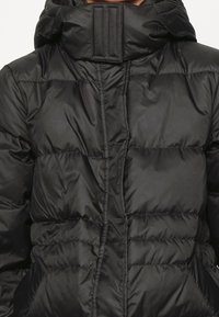 Chaqueta de plumas negra con un alto cuello, con un diseño acolchado, textura brillante y costuras horizontales para aislamiento. Cierre con cremallera.