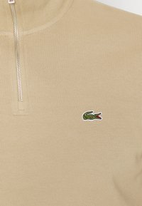 Pulover bej cu fermoar, cu textură cu nervuri, având un mic logo verde de crocodil pe partea stângă a pieptului. Accente cu fermoar argintiu.
