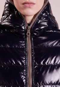 Marinblå, glänsande pufferväst med hög krage, horisontell quiltad textur och en kontrasterande beige dragkedjedetalj. Slät finish.