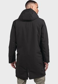 Schwarzer wasserdichter Parka mit Kapuze, der ein elegantes Design, einen langen Schnitt, strukturierte Stoffe und kleine reflektierende Akzente auf dem Rücken aufweist.