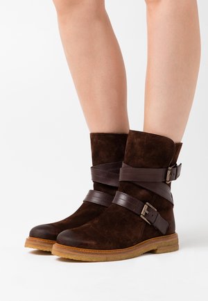 Jambes de femme portant des bottines en daim marron foncé avec des sangles en cuir croisées et des boucles, semelles en caoutchouc marron clair sur fond blanc.