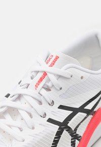 Vit idrottssko med svarta accenter och rosa detaljer. Har en överdel i mesh, rund tå, platta snörningar och en framträdande logotyp på sidan.