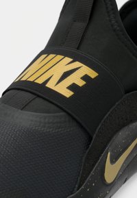 Juodi sportbačiai su tekstūruotu viršumi, auksiniu Nike prekės ženklo užrašu ant dirželio ir taškinė juoda pado tekstūra su auksiniu akcentu.