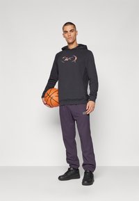 Sort hoodie med et farverigt logo, parret med lilla sweatpants og sorte sneakers. Basketballen holdes i venstre hånd.