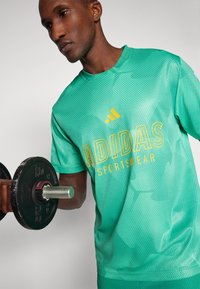 Camisola desportiva verde da Adidas com um padrão texturado, apresentando um logo amarelo e o texto "SPORTSWEAR", usada enquanto levanta um haltere.