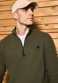 Olivově zelený svetr s relífným zipem, texturovaná tkanina, černý logotyp na hrudi, přeložený přes bílou košili, umístěný na pozadí ze dřeva.