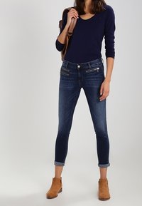 Jeans skiny de mezclilla azul oscuro con puños enrollados, que cuentan con bolsillos con cremallera y un diseño ajustado. Combinados con una camiseta de manga larga en navy.