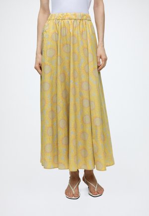 Femme portant une jupe longue jaune à motifs de tournesols, associée à un haut blanc et des sandales beiges.