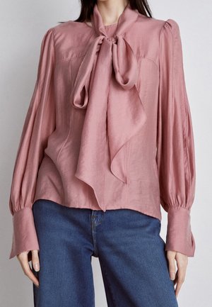 Femme portant un chemisier rose poudré à manches longues avec un grand nœud noué au cou et un jean bleu.