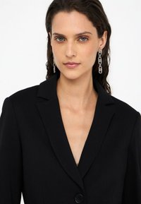 Blazer negro con solapas de pico y cierre de botón único. La modelo lleva pendientes de plata en forma de cadena. Su cabello es ondulado y oscuro.