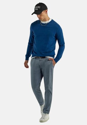 Uomo con maglione blu navy, pantaloni grigio chiaro, sneaker bianche, cappello nero con la scritta "Milano 1984" e collana d'argento, in piedi con una mano in tasca.