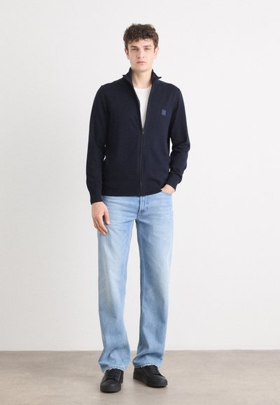 Suéter azul marino con cierre de cremallera en tejido suave, combinado con jeans de un azul claro y zapatillas negras. Presenta un cuello alzado y un logo discreto en el pecho.