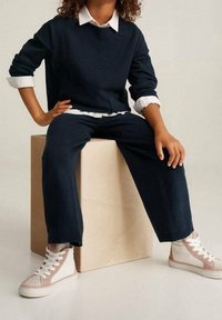 Suéter de punto azul marino con una camisa blanca debajo, pantalones azul marino a juego y zapatillas altas de color rosa claro y blanco, sentado sobre una caja de madera.