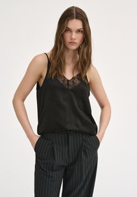 Top in raso nero con dettagli in pizzo lungo il decolleté, abbinato a pantaloni a righe scure. Texture morbida, spalline sottili regolabili.