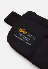 Fekete nylon tasak, textúrált felülettel, kiemelkedő sárga és fehér márkacímkével, és egy fekete pánttal a tetején.