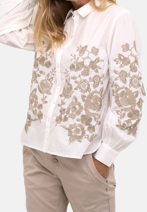 Hvid skjorte med knapper og beige blomsterbroderi på fronten og ærmerne, kombineret med beige bukser, model med hånden i lommen.