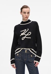 Schwarzer gerippter Strickpullover mit cremefarbenen Akzenten, hohem Kragen und großem gesticktem Logo. Kombiniert mit hochgeschnittenen dunklen Jeans.