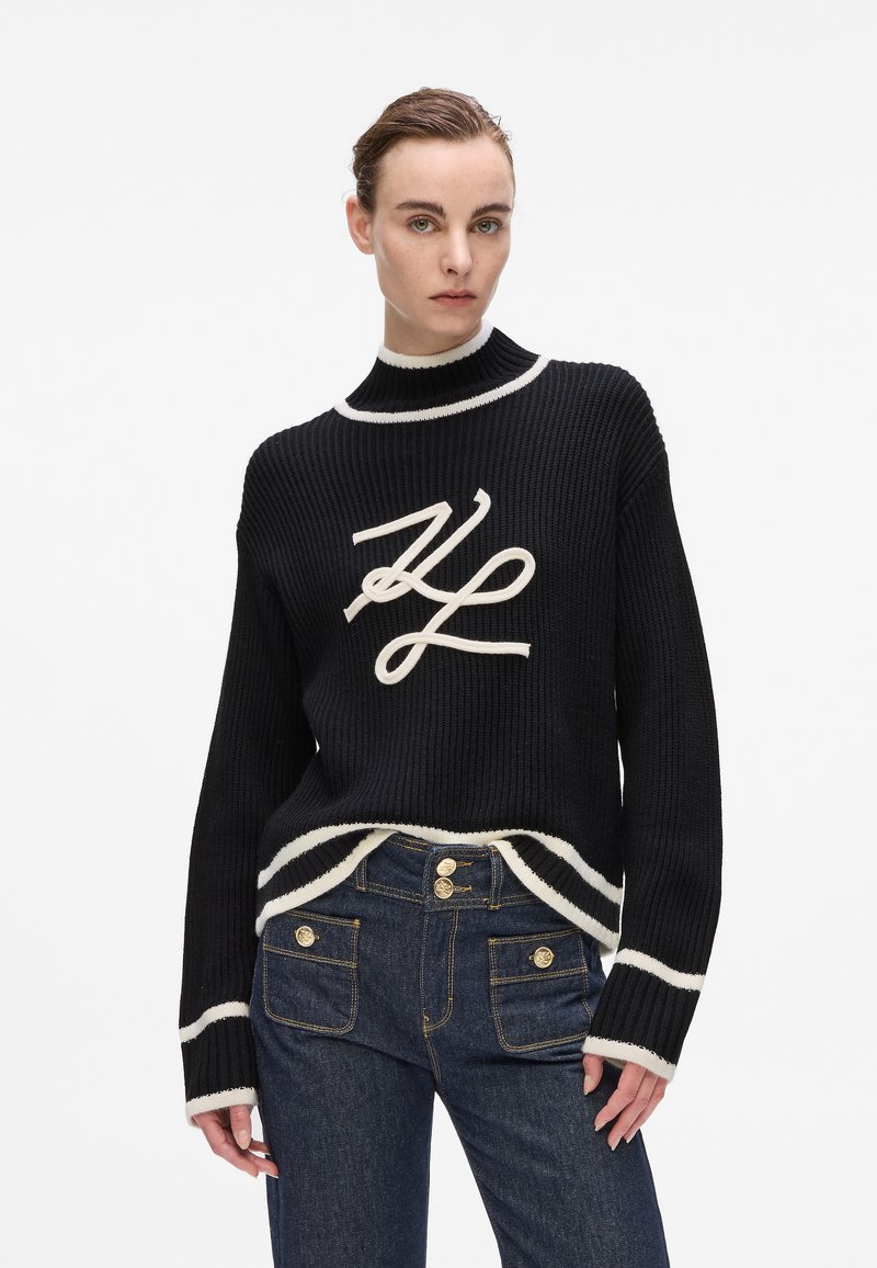 Schwarzer gerippter Strickpullover mit cremefarbenen Akzenten, hohem Kragen und großem gesticktem Logo. Kombiniert mit hochgeschnittenen dunklen Jeans.