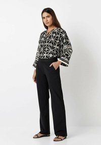 Schwarz-weiß gemusterte Bluse mit Glockenärmeln und einem Schlüsselloch-Ausschnitt, kombiniert mit lockeren schwarzen Hosen und flachen Sandalen.